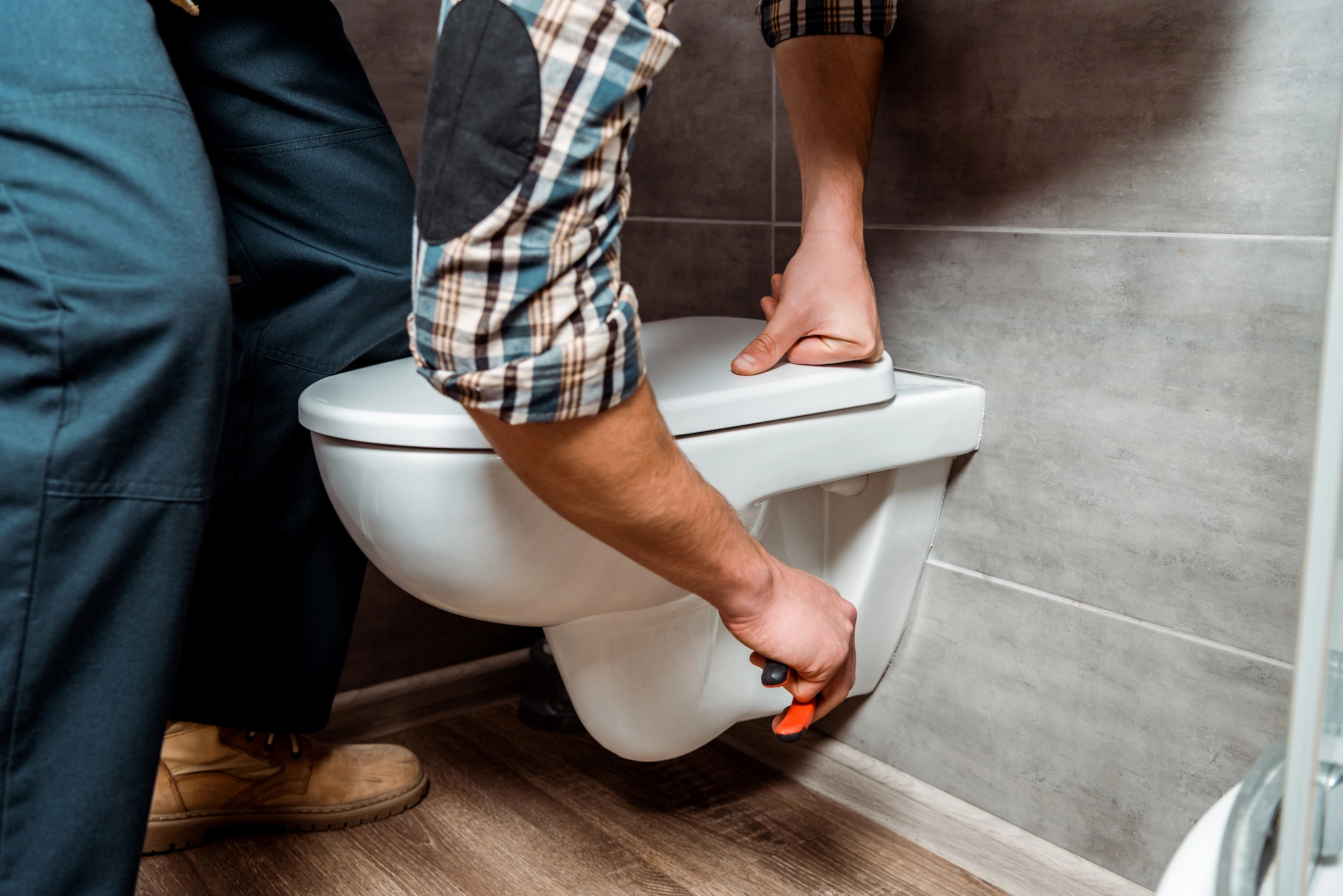A person wearing a plaid shirt and work boots is seen fixing a wall-mounted toilet with a wrench. The bathroom has gray tiled walls and a wooden floor.