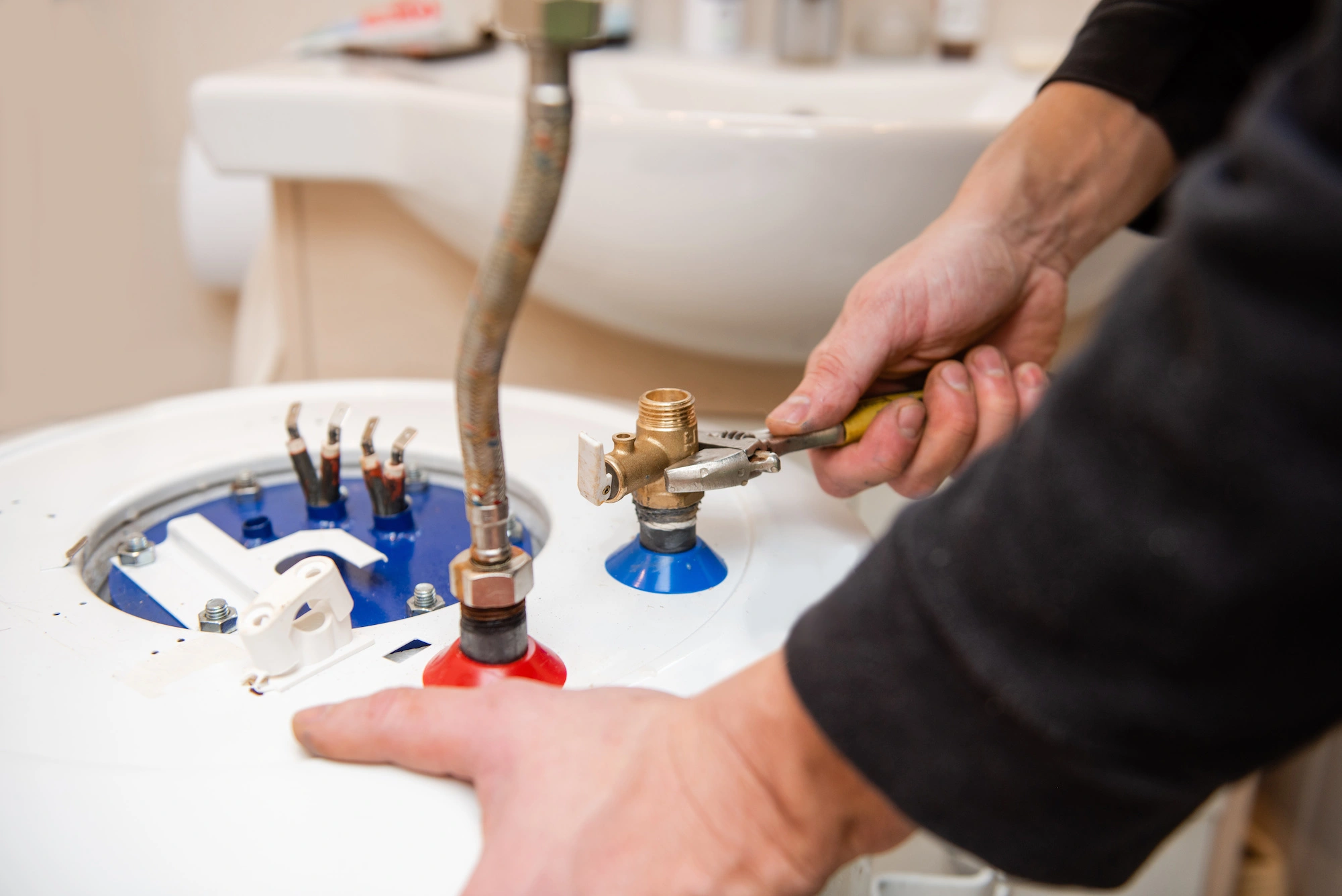 A person is using a wrench to adjust a component on the top of a water heater. Various pipes and connections are visible, and the setting appears to be a home utility area with a sink in the background.