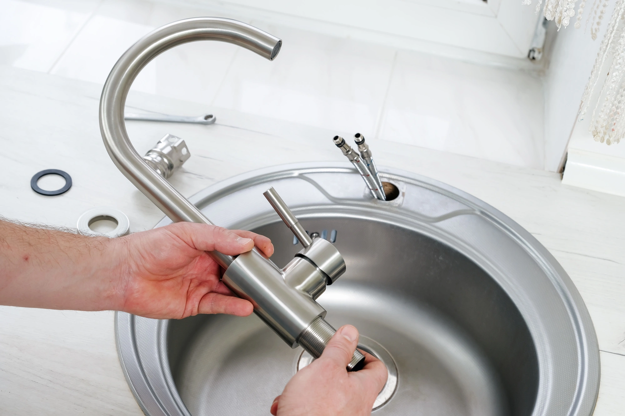 Person's hands installing a metal kitchen faucet over a circular stainless steel sink. Various installation tools and parts are visible on the countertop nearby.