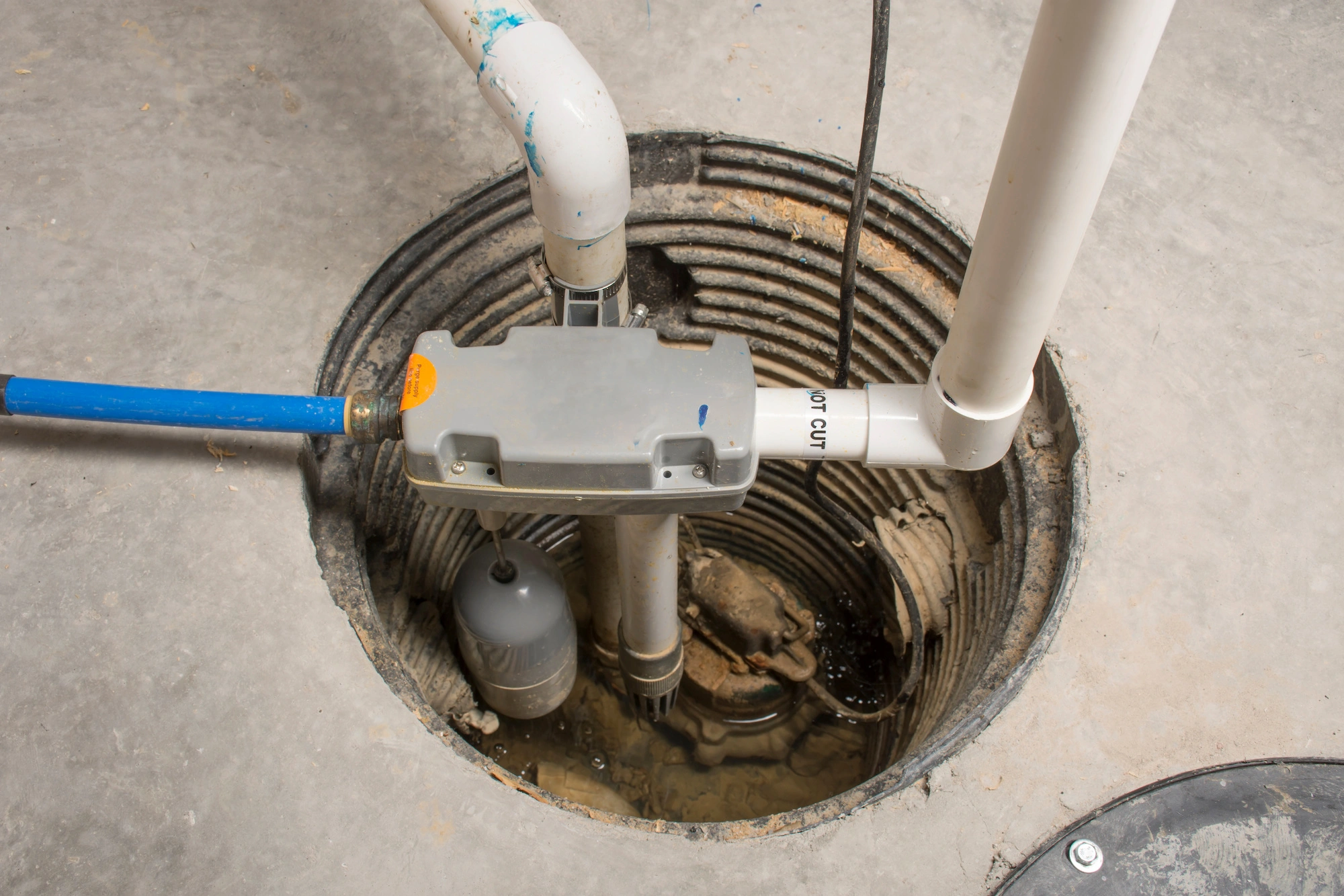 A sump pump system installed in a basement pit, with visible pipes and a water-filled hole. The setup includes a float switch and various PVC connections for directing water flow. The surrounding floor is concrete.