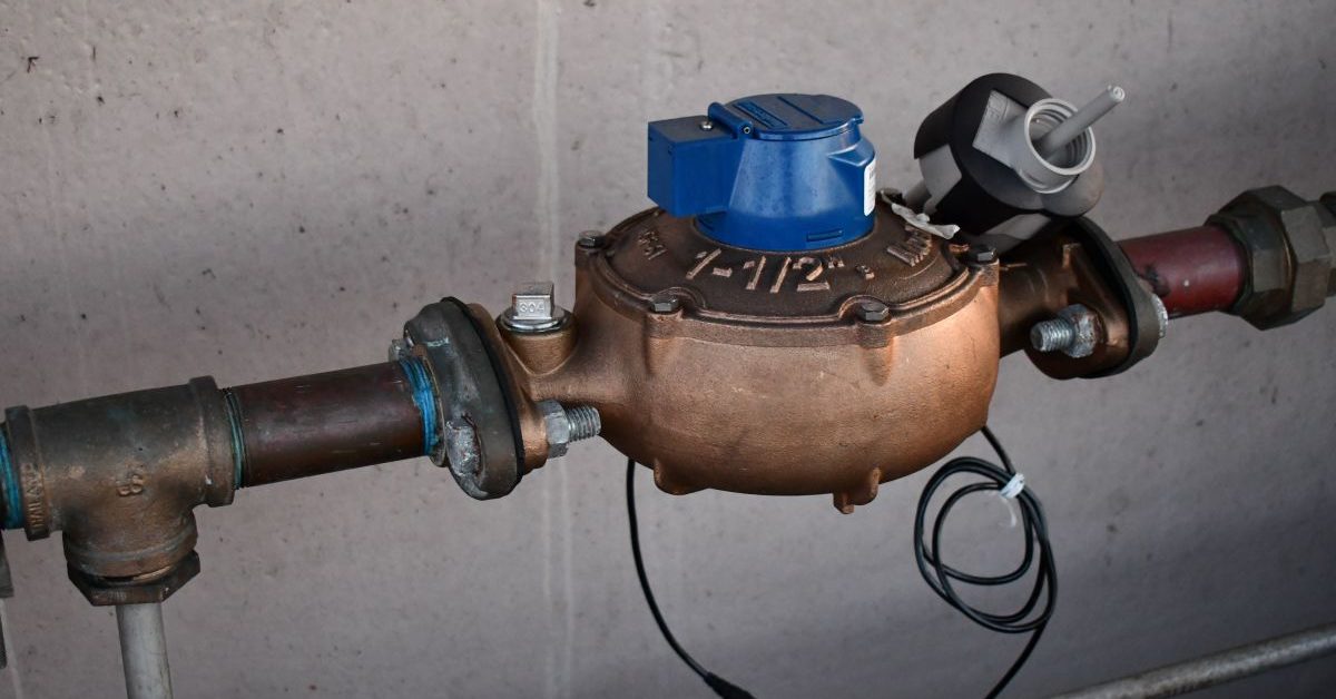 A close-up of a bronze water meter with a blue digital reading device attached, connected to metal pipes against a concrete wall.