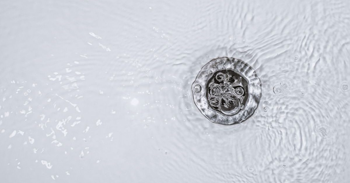 Close-up of water swirling around a metal drain in a white sink, creating ripples and small bubbles on the surface.